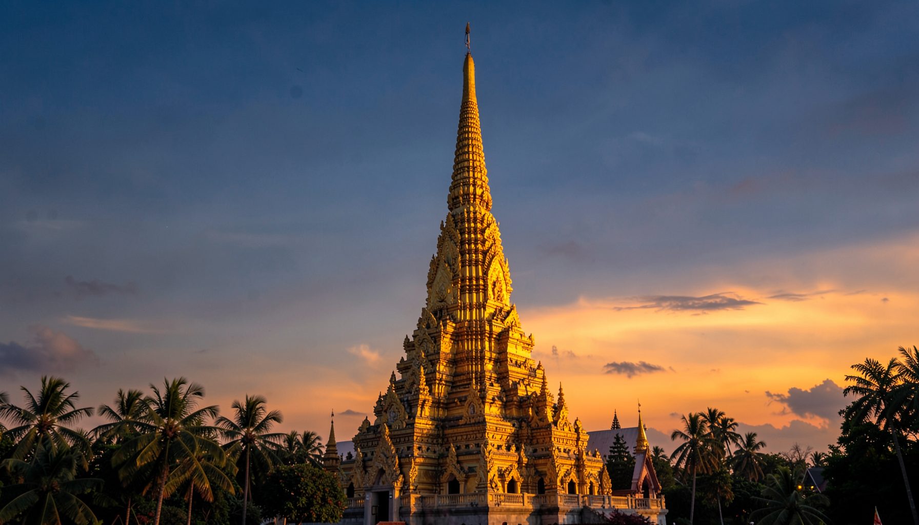 Tall golden ornate pyramid-shaped tower silhouetted against a dramatic orange and blue sunset sky, surrounded by palm trees
