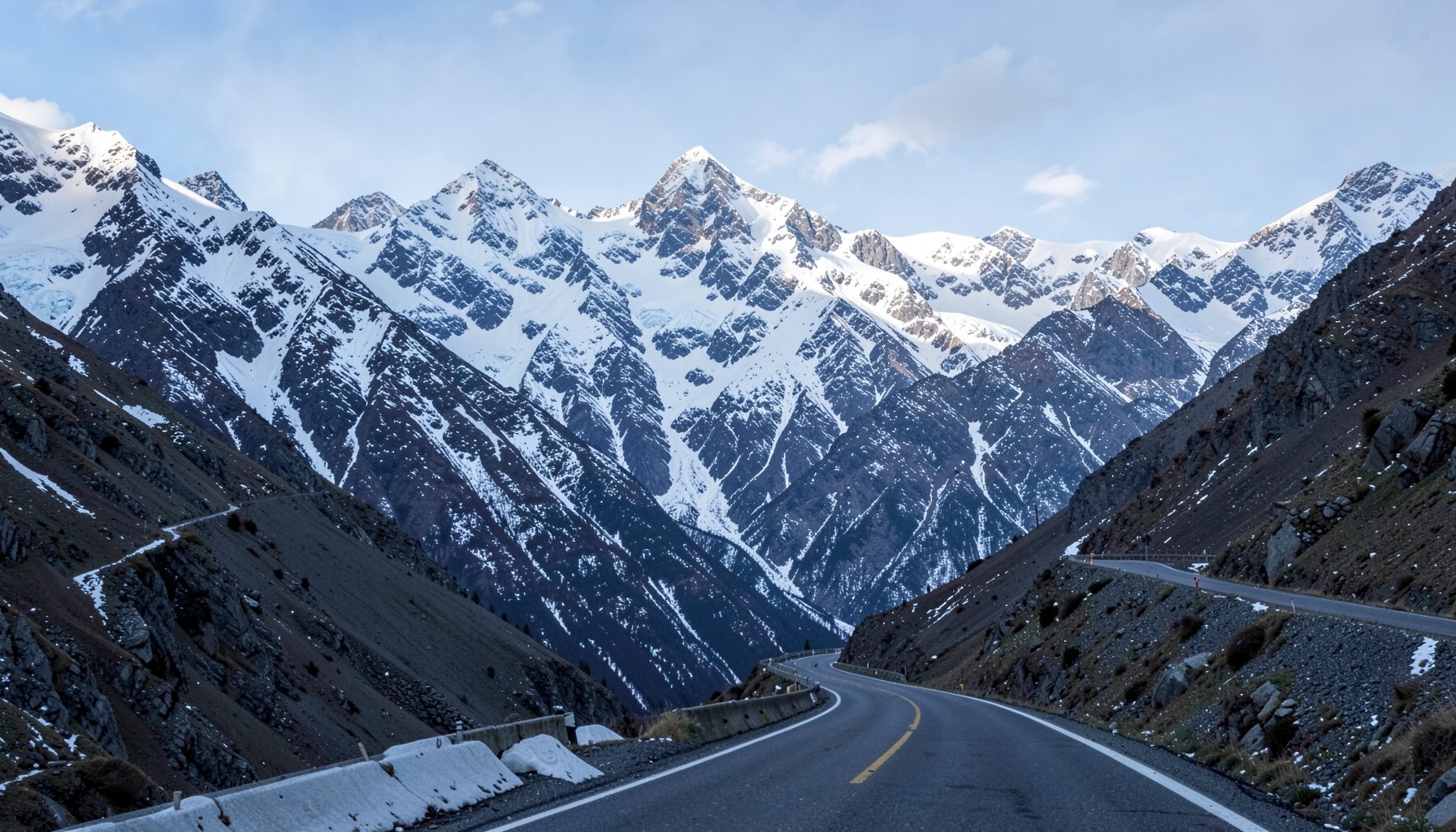 Winding mountain road leading toward snow-capped peaks, gray asphalt against dark rock and white snow