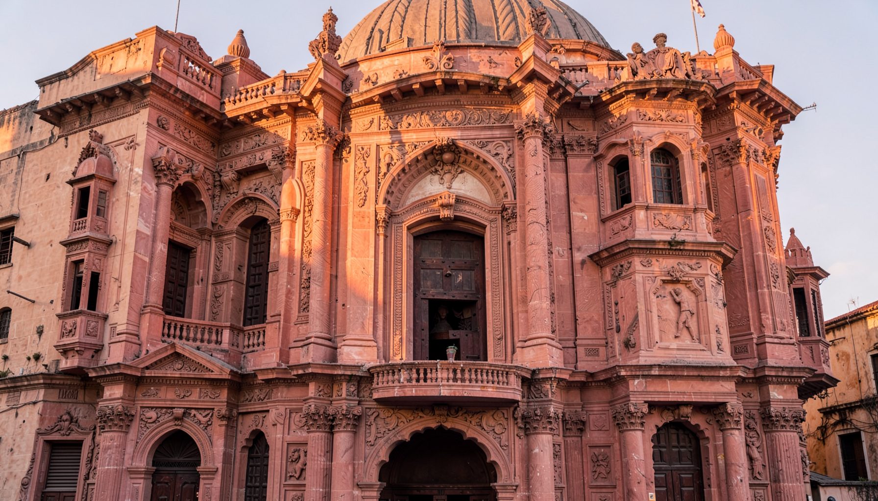 Ornate historic building facade with domed roof and carved stone details, warm pink and terracotta tones under a pale sky