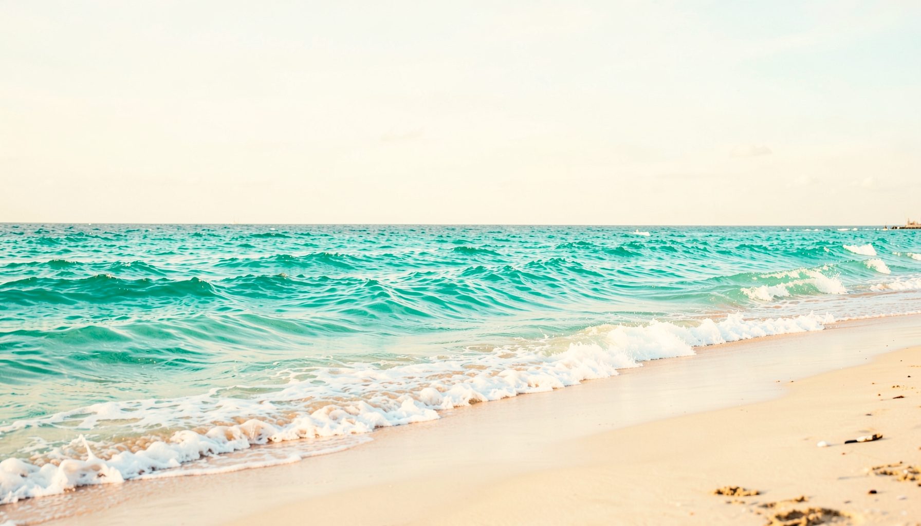 Ocean waves rolling onto a sandy beach, turquoise water and white foam under a pale sky